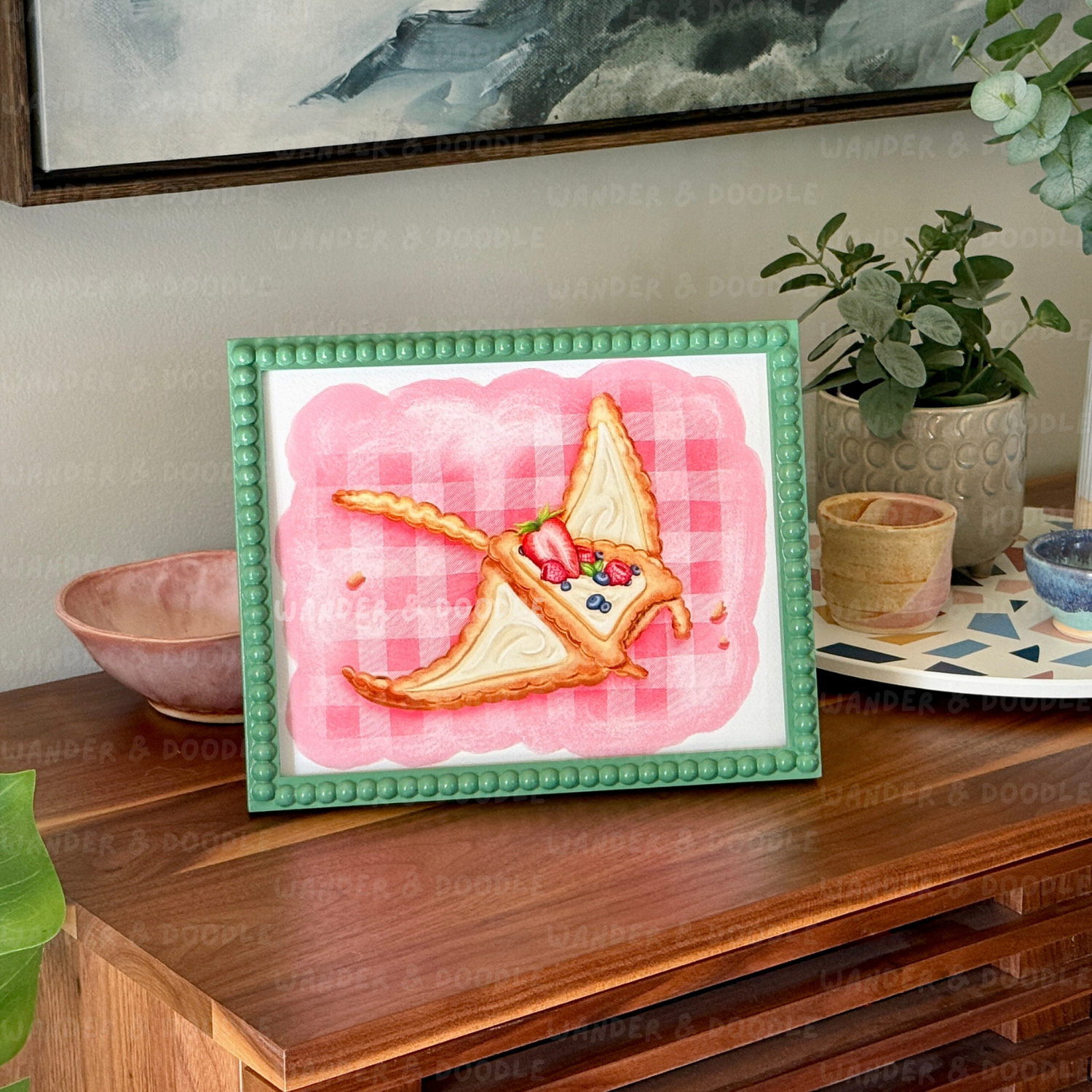 Framed artwork of a slice of pie in the shape of a manta ray with toppings on a pink checkered background, displayed on a wooden surface.