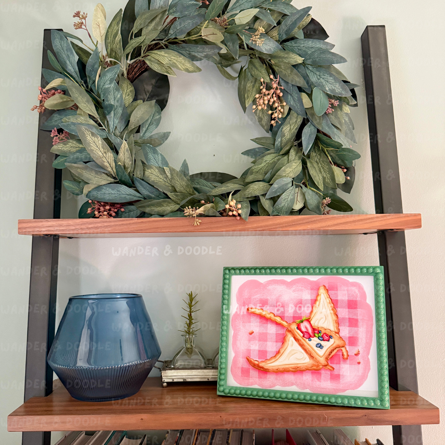 Wooden shelf with a green leafy wreath, blue vase, and framed artwork on a white wall. The art work is fruit tart in the shape of a manta ray