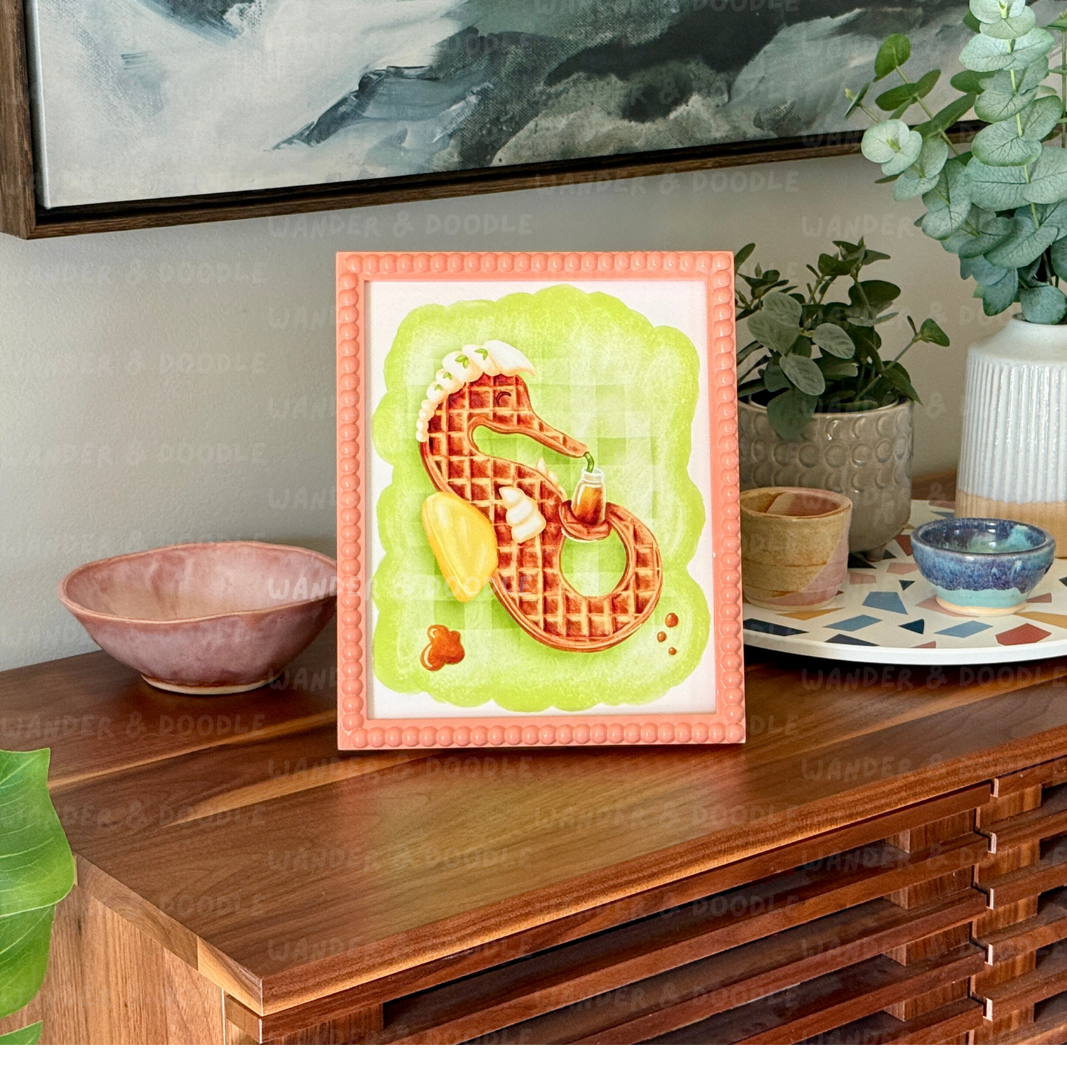 Framed artwork of a waffle in the shape of a seahorse on a wooden surface with decorative items.