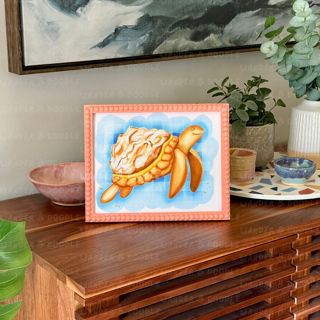 Framed artwork of a turtle  in the shape of a lemon meringue pie  on a wooden surface with decorative items.