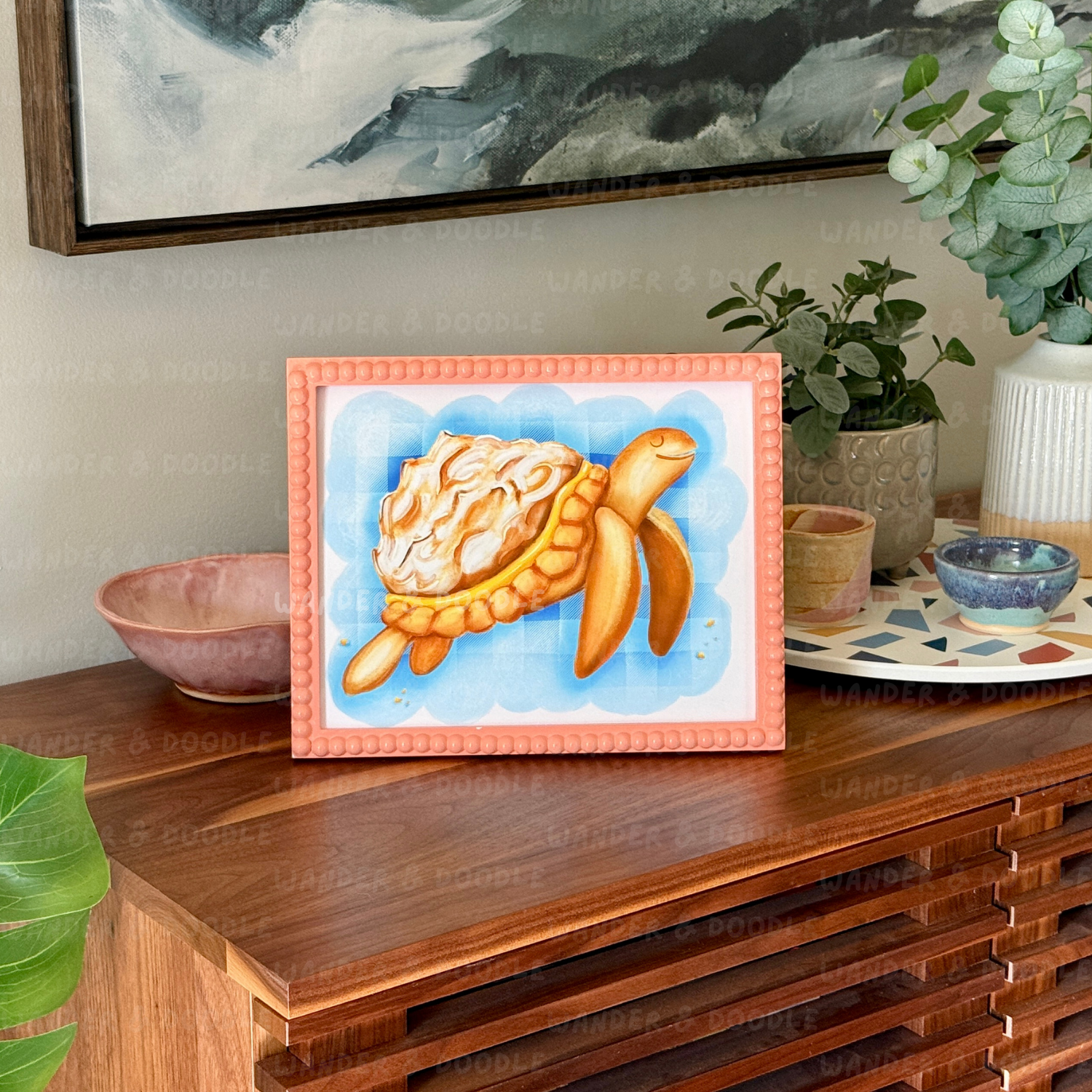 Framed artwork of a turtle  in the shape of a lemon meringue pie  on a wooden surface with decorative items.