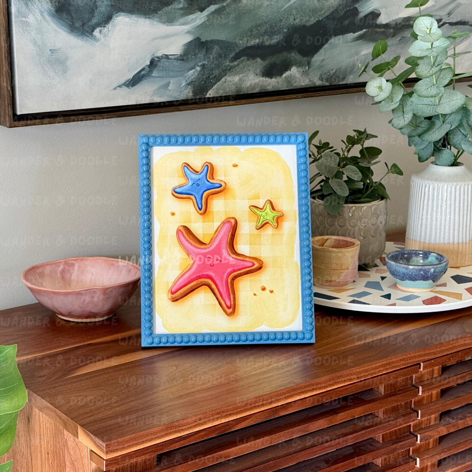 Framed artwork of colorful starfish sugar cookie illustration on a wooden surface with decorative items.